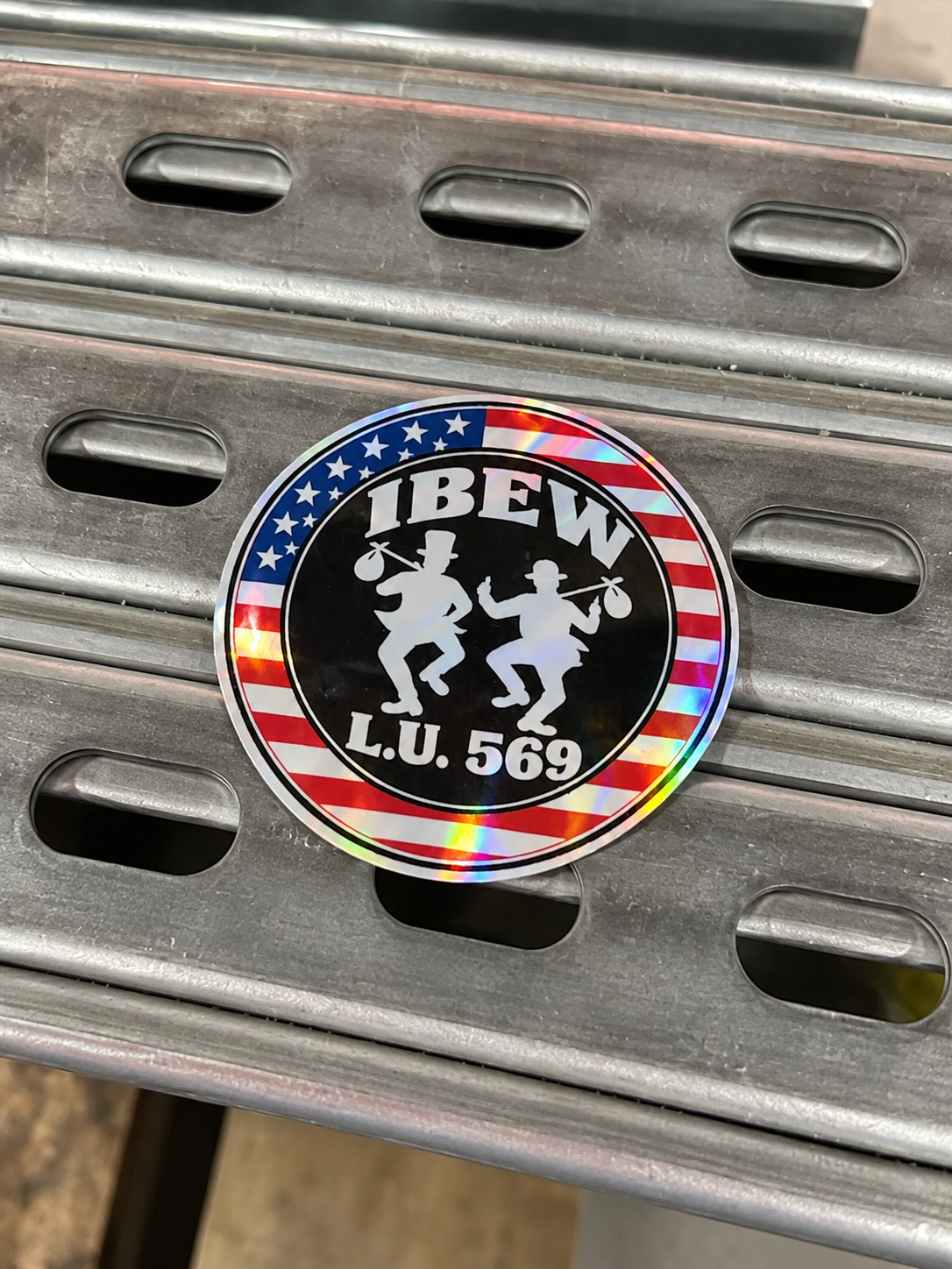 Enhanced product photo of holographic IBEW Traveler Tramps decal on steel Unistrut; L.U. 569 shown as a personalization example